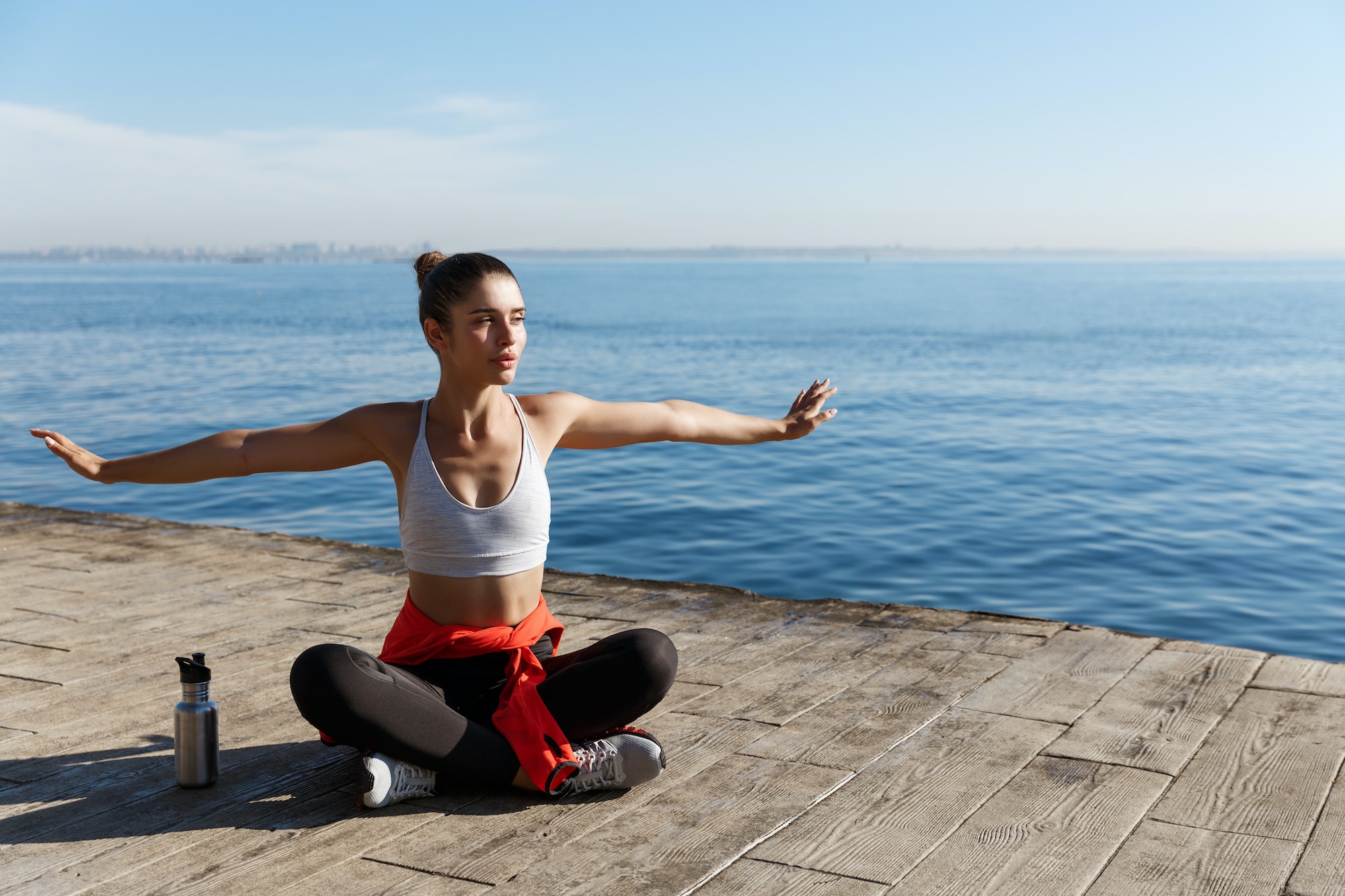 Outdoor shot of relaxed young woman sitting on wooden pier and looking at the sea, doing yoga
