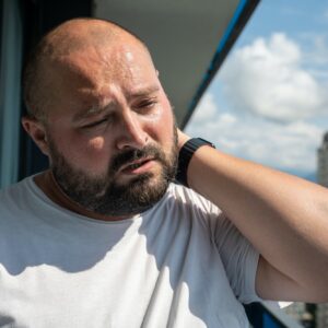 Bearded man in summer sun on balcony. Guy closed eyes in displeasure discomfort holding on to neck,